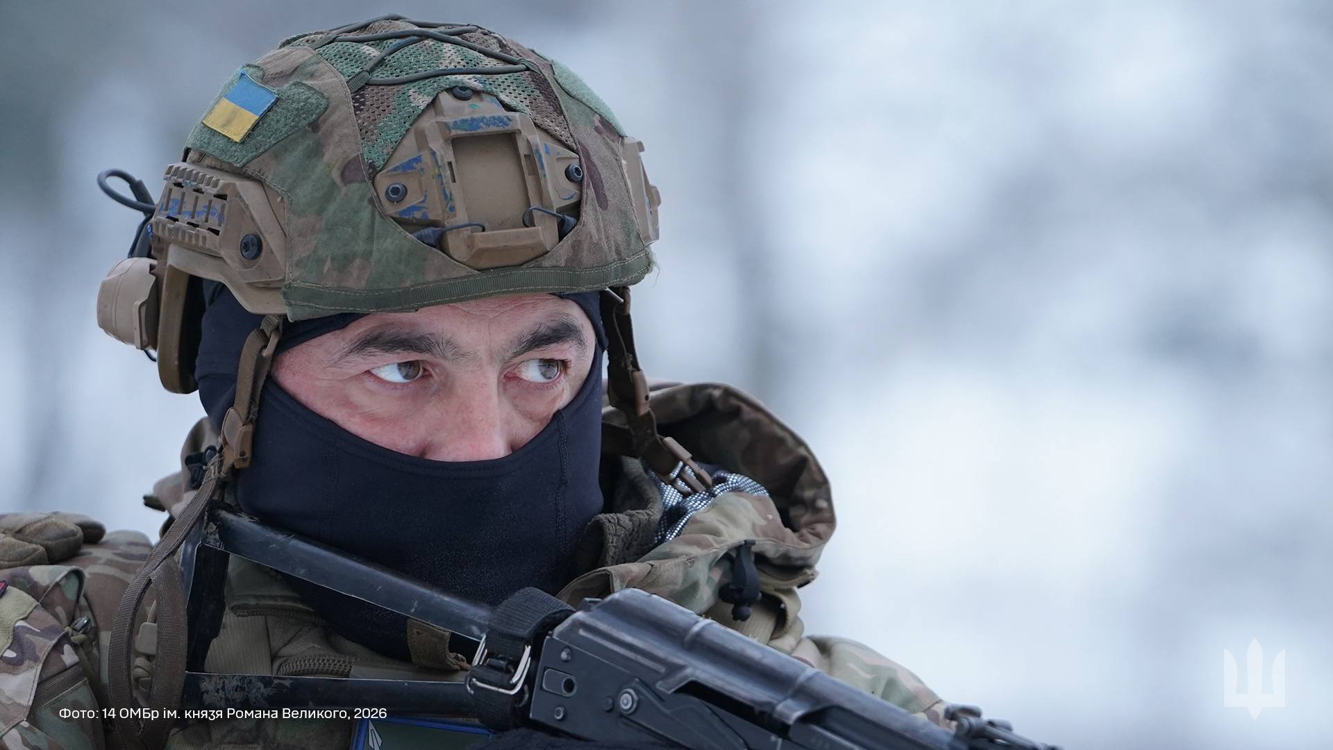 Український військовий у бойовому спорядженні: у шоломі з шевроном, балаклаві та з автоматом напоготові. Його зосереджений погляд убік і зимовий фон підкреслюють напружену обстановку та готовність до дії.