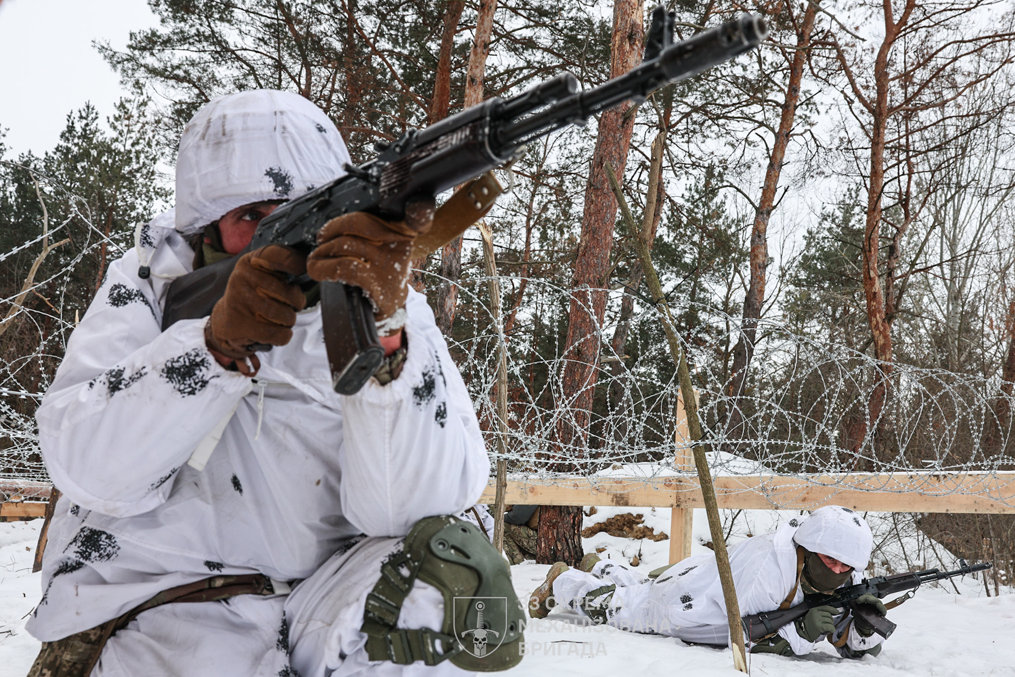 Українські військові в зимовому маскуванні, які займають бойові позиції в лісі, вкритому снігом. Один боєць веде спостереження й тримає автомат напоготові, тоді як інший лежить нижче, прикриваючи напрямок уздовж колючого дроту. Сцена передає напружений момент бойового чергування в холодних зимових умовах.