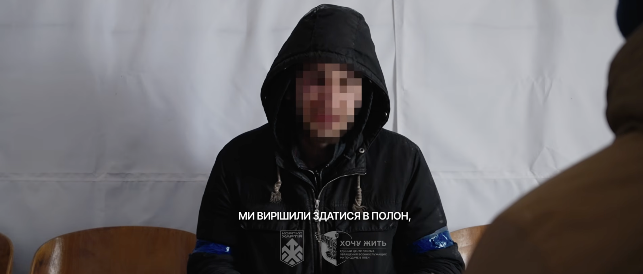 На фото видно людину в темній куртці з капюшоном, обличчя якої навмисно розмите, вона сидить на стільці в приміщенні з білим тлом. На екрані поверх зображення є напис українською про рішення здатися в полон, а на рукаві помітна синя стрічка.