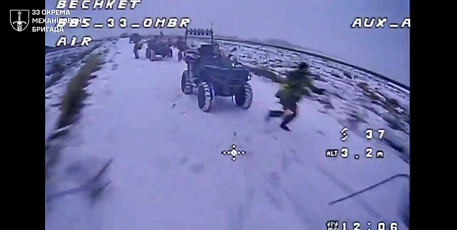 Група російських військових рухається засніженою дорогою на кількох квадроциклах. Один із них біжить поруч із технікою, тоді як інші зупинилися або готуються продовжити рух. Кадр має характерні елементи відео з дрона: прицільну мітку, телеметрію та холодну, приглушену кольорову гаму.