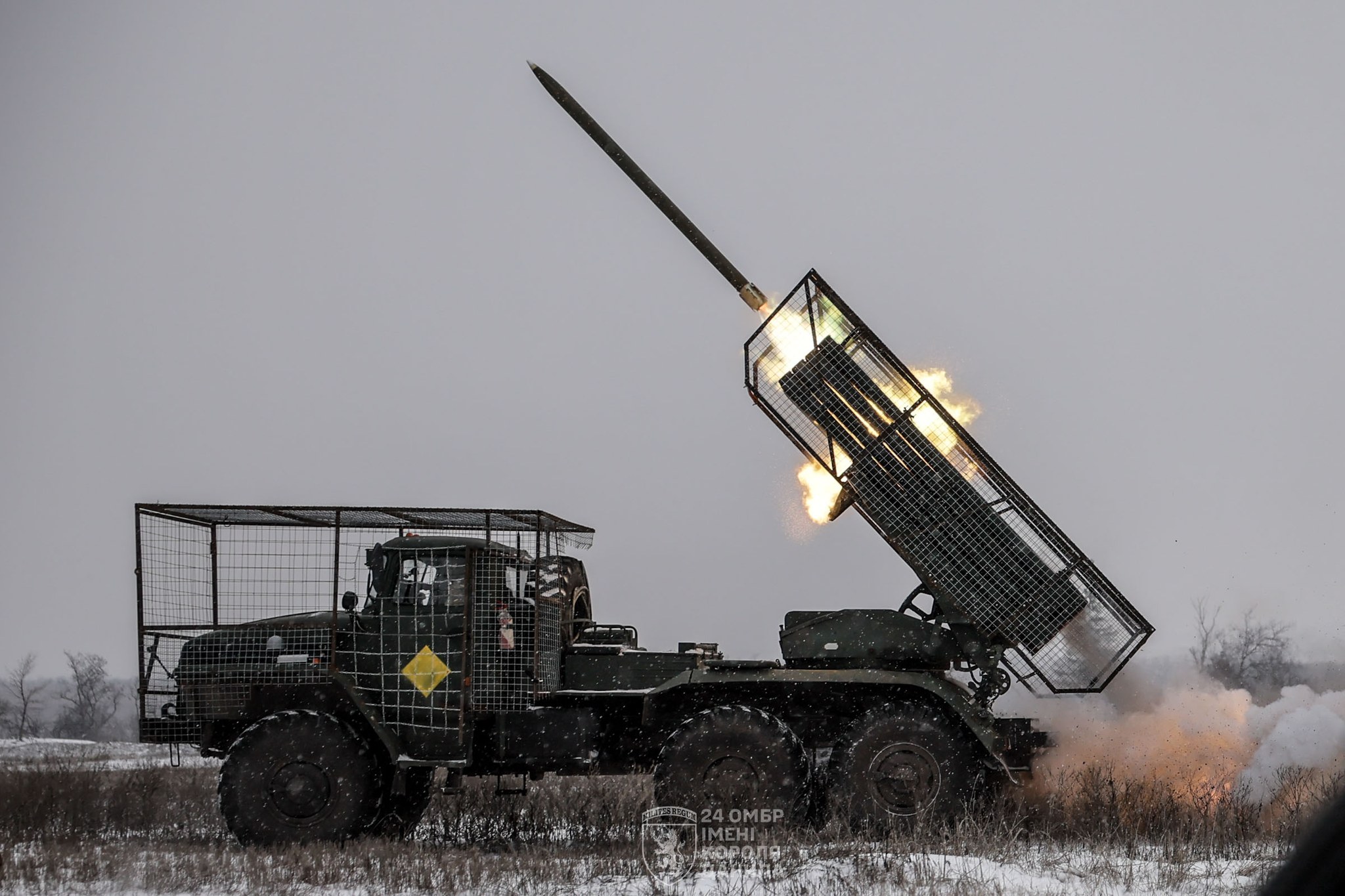 На засніженому полі військова вантажівка з реактивною пусковою установкою в захисній металевій клітці здійснює запуск ракети. У момент пострілу видно полум’я й дим, а навколо — зимовий пейзаж із низькою рослинністю та сірим небом.
