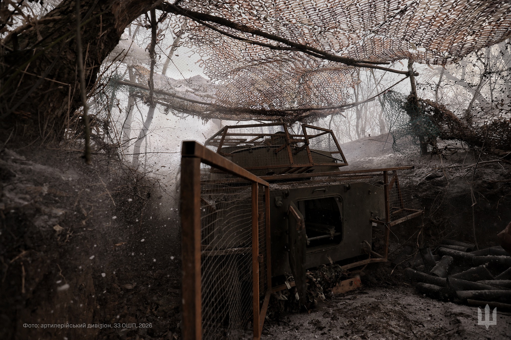 Самохідна артилерійська установка розташована в укритті серед дерев і замаскована сітками та захисними конструкціями. У момент пострілу навколо здіймаються дим, пил і грудки землі, що підкреслює інтенсивність бойової роботи на позиції.