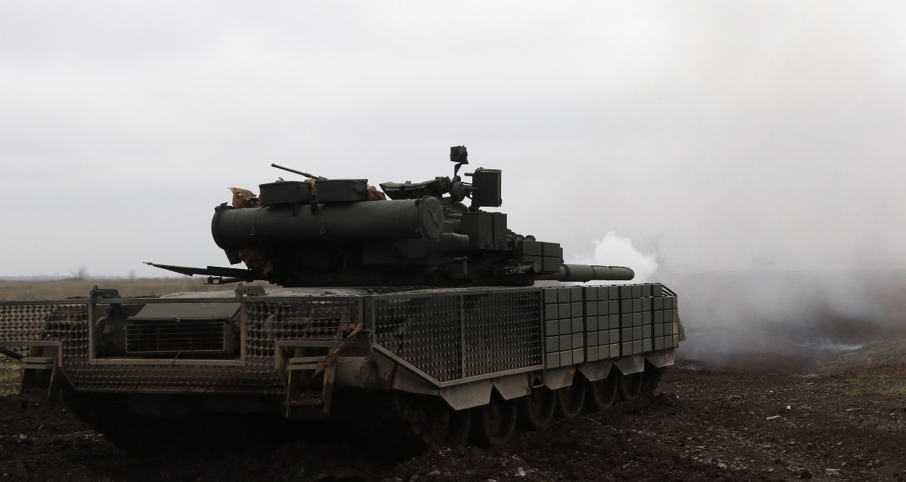 Танк із додатковим захистом на корпусі, який рухається по розмитому польовому ґрунту в туманну погоду. Зі ствола підіймається дим, що свідчить про нещодавній постріл або роботу двигуна під час бойових дій.