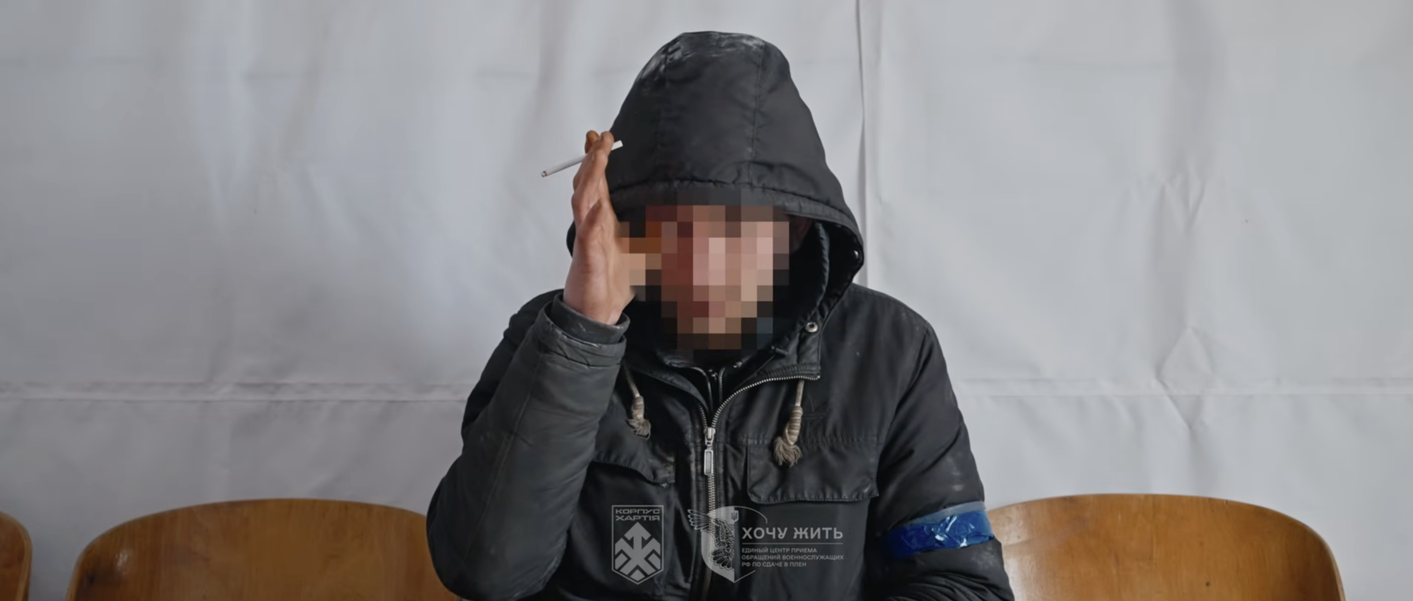 Чоловік у темній куртці з капюшоном сидить на стільці перед світлим тлом, його обличчя замасковане пікселізацією. Він тримає біля скроні руку з сигаретою, а на рукаві видно синю пов’язку та нашивку з написом «Хочу жить».