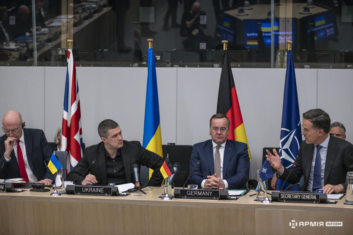 Представники України, Німеччини та міжнародних структур сидять поруч за столом переговорів, уважно слухаючи виступ співрозмовника. Прапори позаду та таблички з назвами країн підкреслюють офіційний характер дипломатичної дискусії.