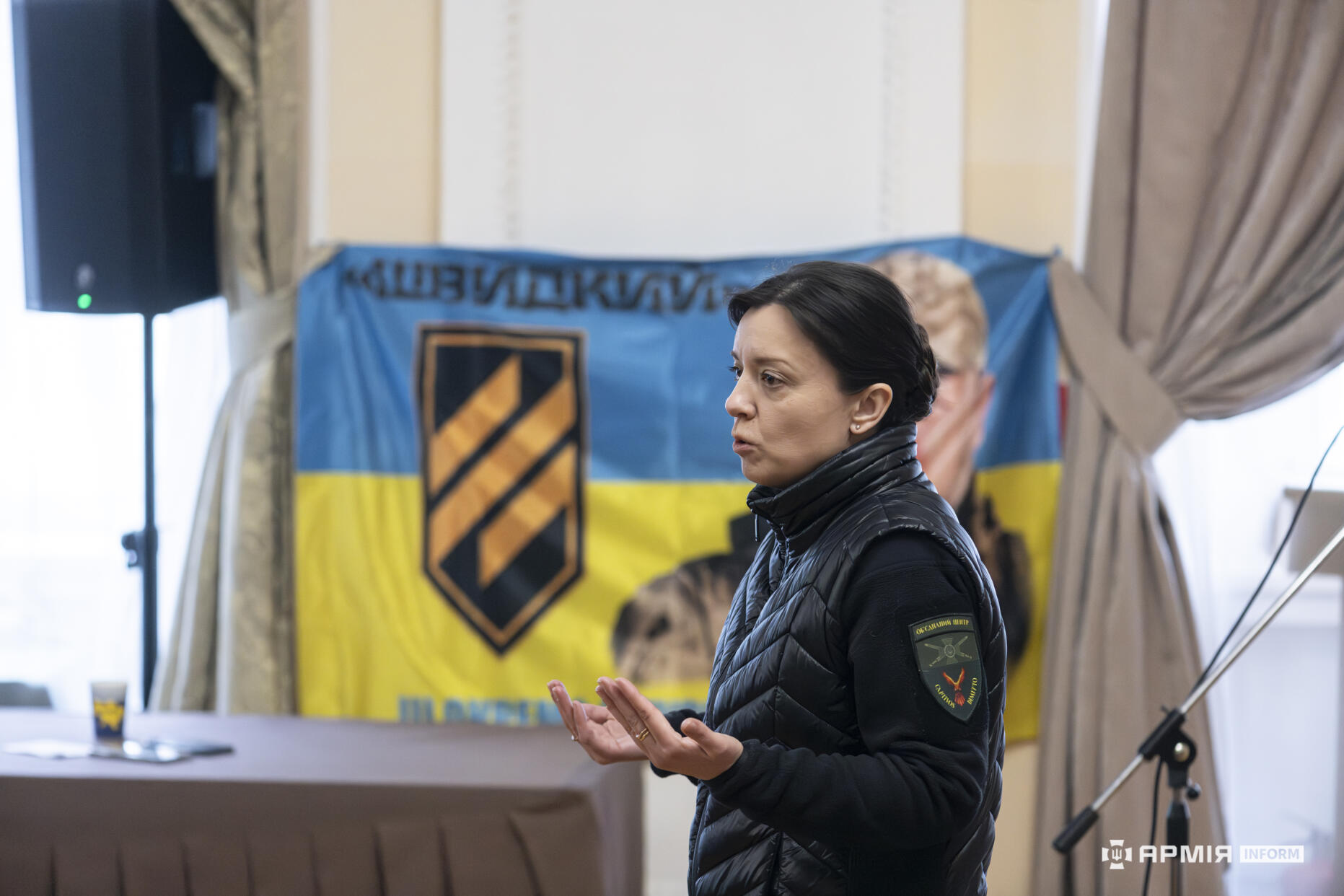 Жінка виступає перед аудиторією у залі, жестикулюючи під час пояснення. Позаду неї розміщений синьо-жовтий банер із символікою військового підрозділу та зображенням людини.