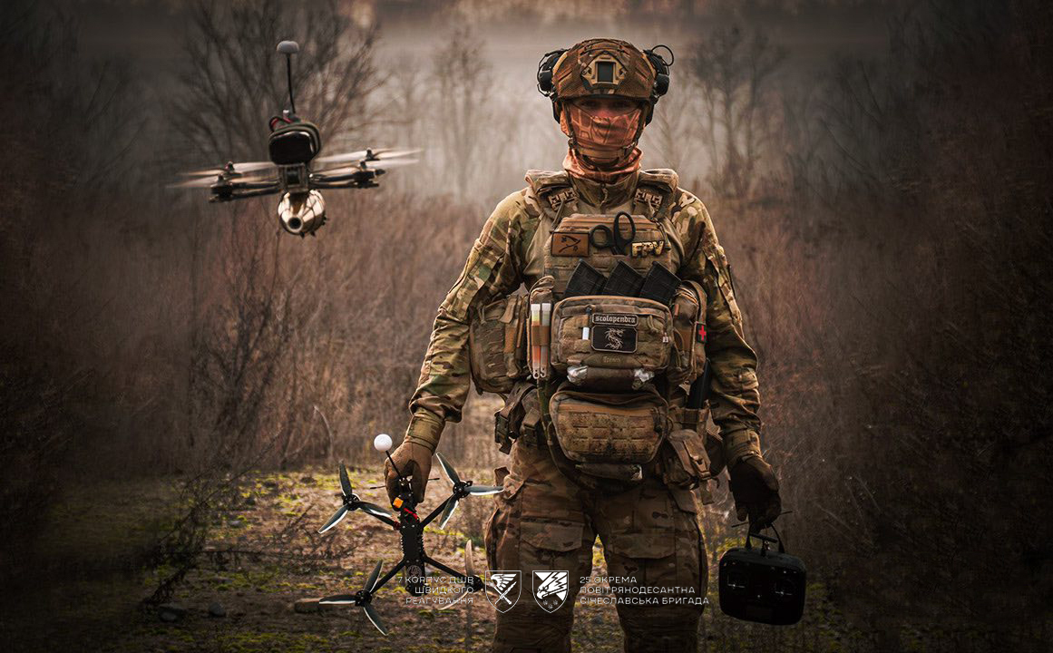 Боєць у повному спорядженні стоїть на тлі лісу, тримаючи FPV-дрон і пульт керування, тоді як інший безпілотник зависає позаду нього. Його екіпірування та поза підкреслюють готовність до виконання бойових завдань із використанням сучасних технологій.