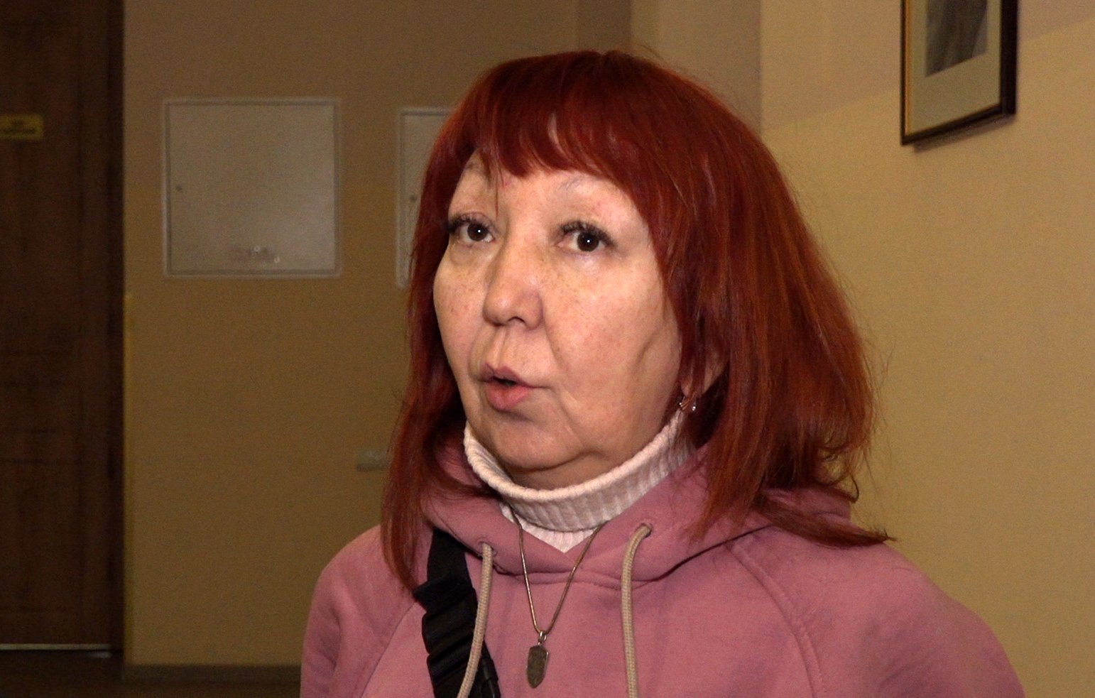 Жінка з рудим волоссям у рожевій кофті дає коментар у приміщенні, звертаючись до співрозмовника поза кадром. Її обличчя виражає зосередженість і серйозне ставлення до теми розмови