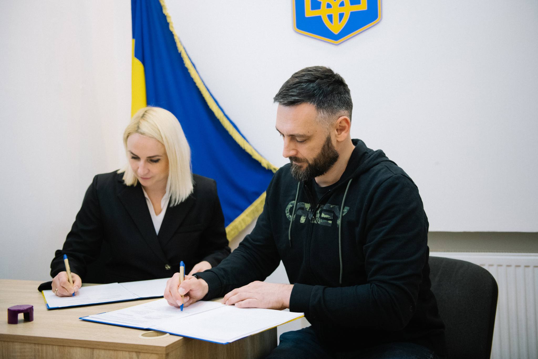 Жінка та чоловік сидять за столом і підписують документи, використовуючи сині ручки. Позаду них видно український прапор і герб, що підкреслює офіційний характер зустрічі.