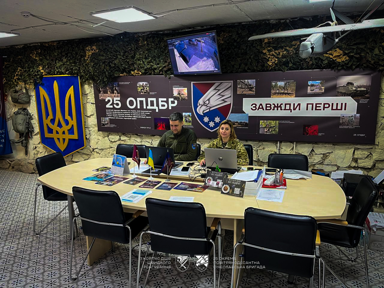 У приміщенні рекрутингового центру двоє військових працюють за ноутбуками за столом із інформаційними матеріалами. На стінах розміщені символіка бригади, банери та фото, що презентують діяльність підрозділу.