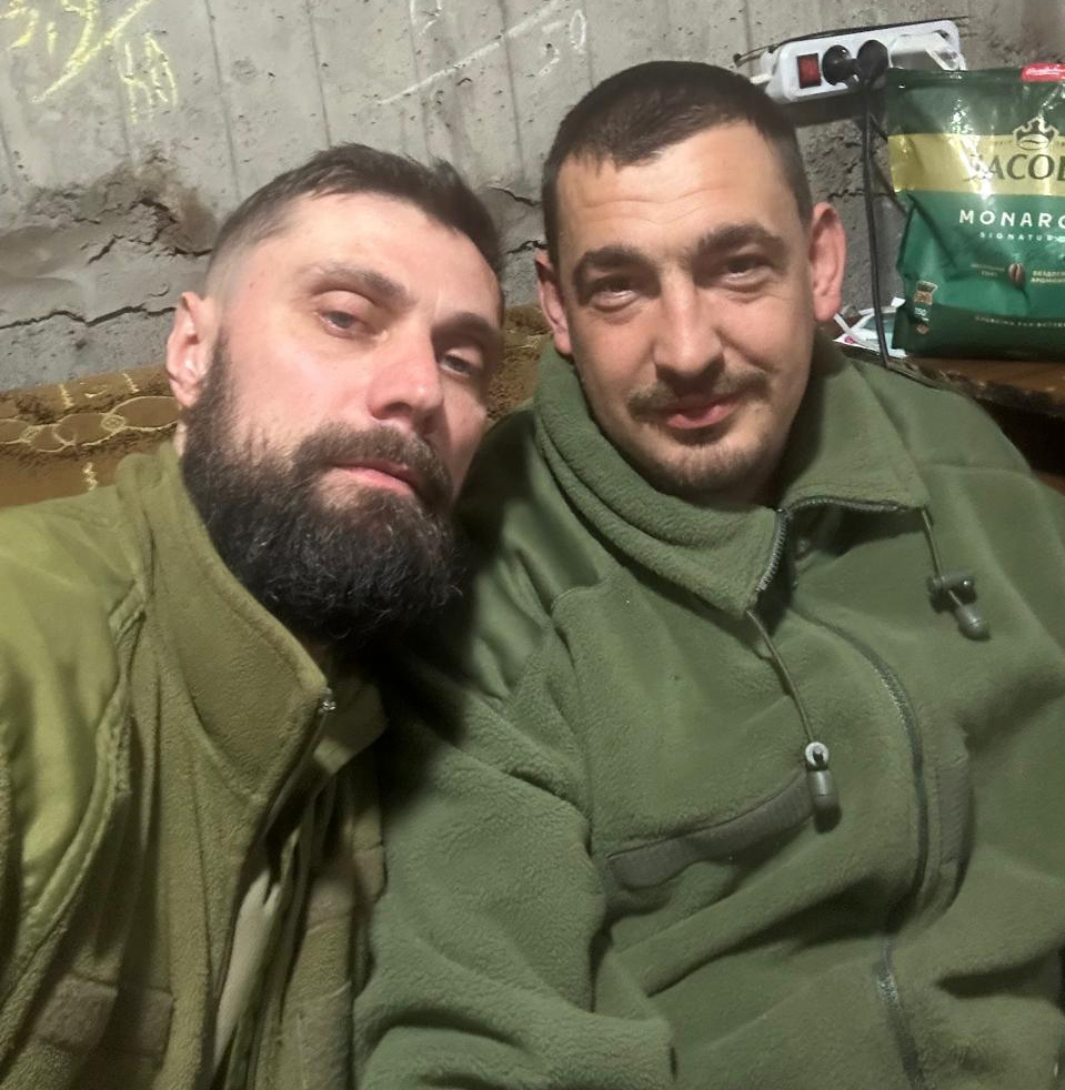 Двоє чоловіків у військовому одязі роблять спільне селфі в приміщенні з бетонними стінами. Вони сидять поруч і дивляться в камеру, відпочиваючи у невимушеній атмосфері.