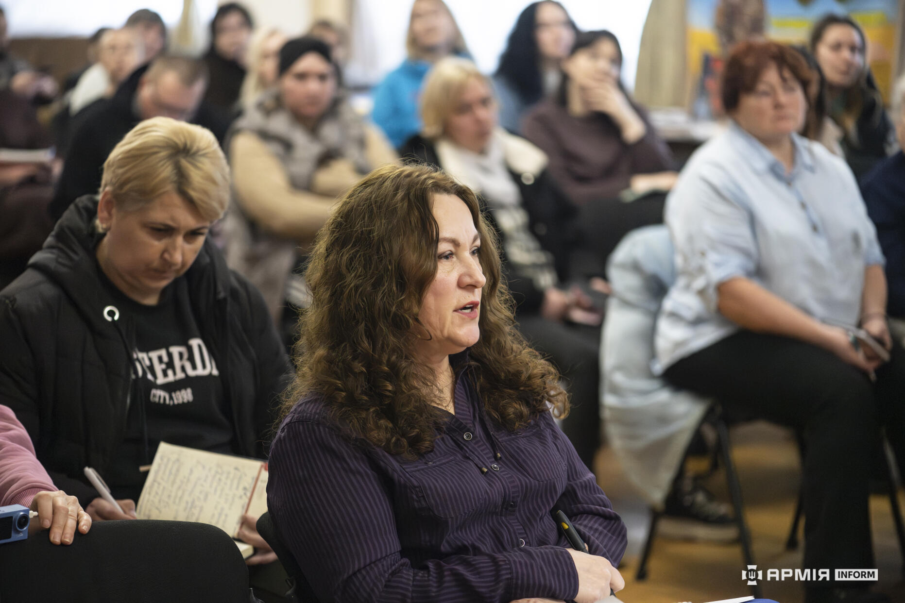 Учасники заходу сидять у залі та уважно слухають виступ, частина з них робить записи в блокнотах. На передньому плані жінка з кучерявим волоссям уважно стежить за обговоренням.