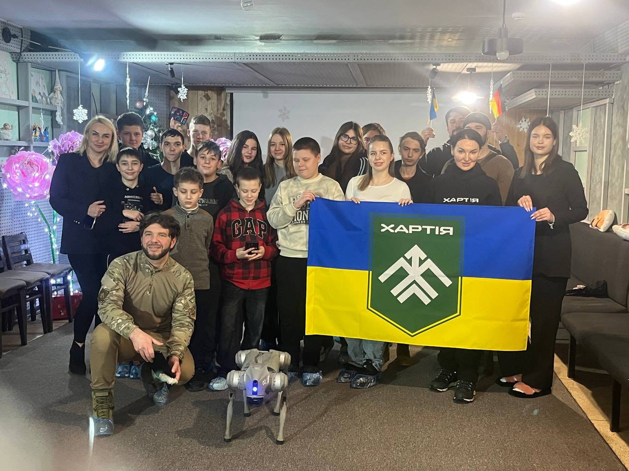 Група підлітків і дорослих позує для спільного фото в приміщенні, прикрашеному святковими елементами. Попереду стоїть невеликий робот-собака, а учасники тримають синьо-жовтий прапор із написом «ХАРТІЯ».