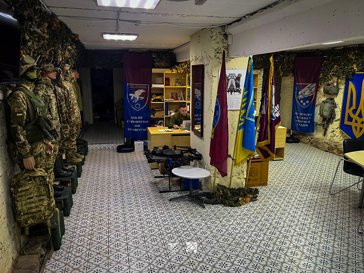 У кімнаті з військовою символікою стоять манекени у формі, а поруч видно обладнання та робоче місце з комп’ютером. Простір оформлений як демонстраційна та робоча зона для ознайомлення з підрозділом.