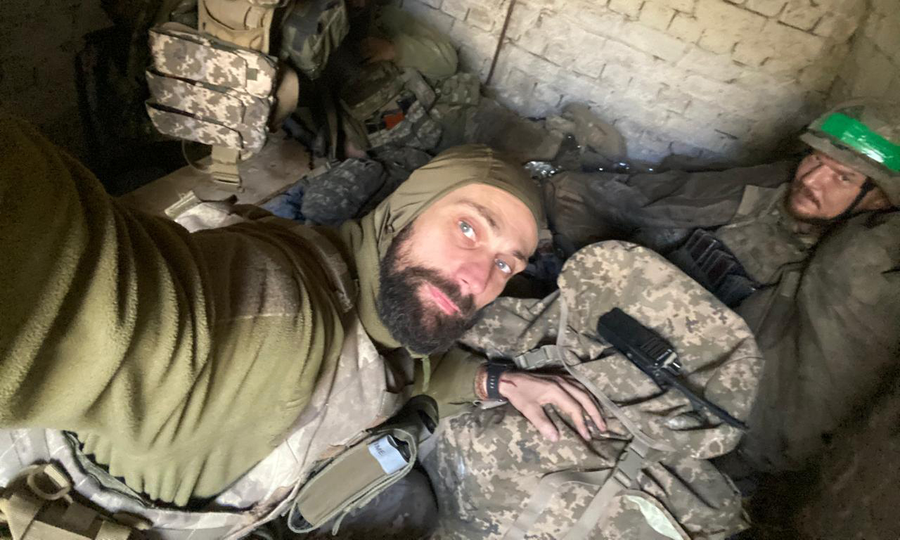 Боєць робить селфі у бліндажі разом із побратимами, які відпочивають на імпровізованих місцях для сну. Поруч видно військове спорядження та зброю, що передає атмосферу фронтового побуту.