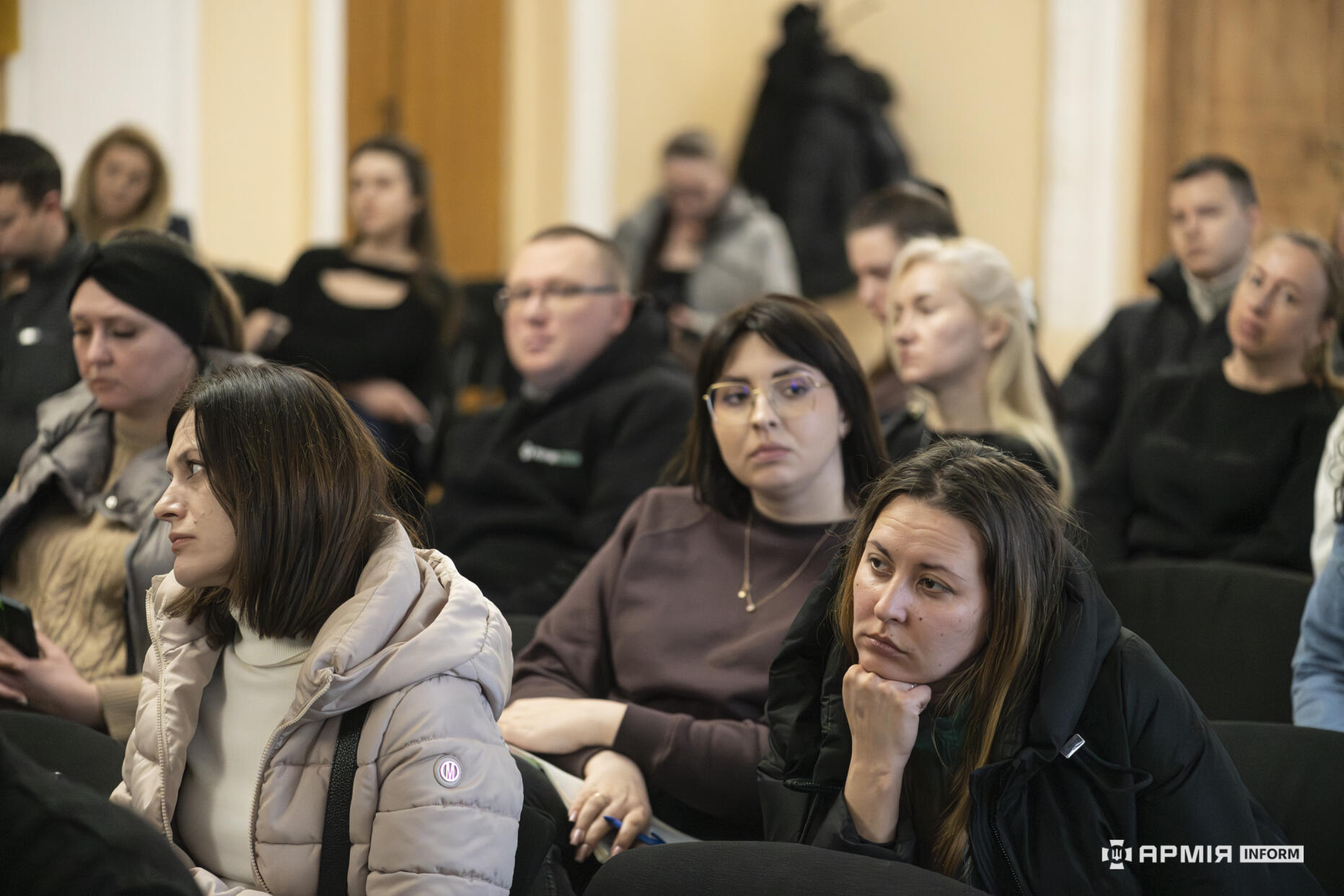 Учасники заходу сидять у залі та уважно слухають виступ, деякі роблять нотатки. Вирази їхніх облич свідчать про зосередженість і зацікавленість у темі обговорення.