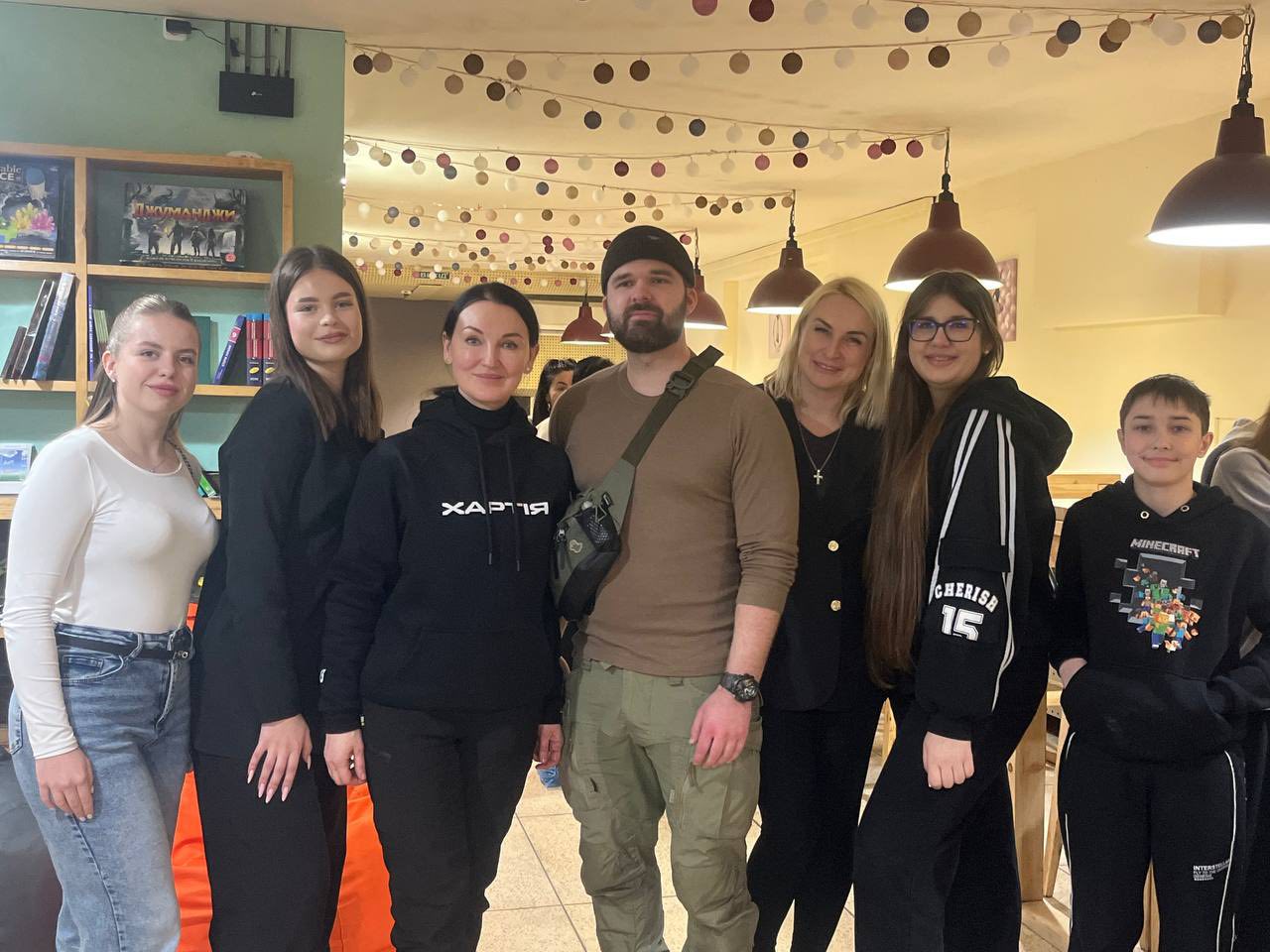 Кілька дорослих і підлітків стоять разом у приміщенні з книжковими полицями та декоративними гірляндами. У центрі групи — чоловік у військовому стилі одягу та дві жінки, одна з яких одягнена у худі з написом «ХАРТІЯ».