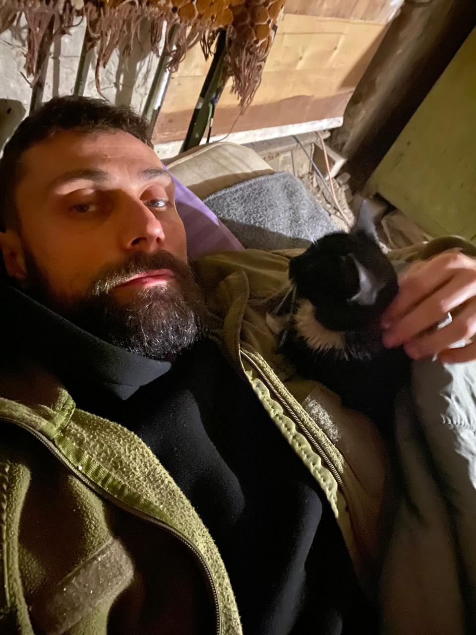Чоловік лежить у приміщенні та тримає на руках чорно-білого кота, якого лагідно гладить. Тварина спокійно сидить поруч, додаючи кадру відчуття домашнього затишку.