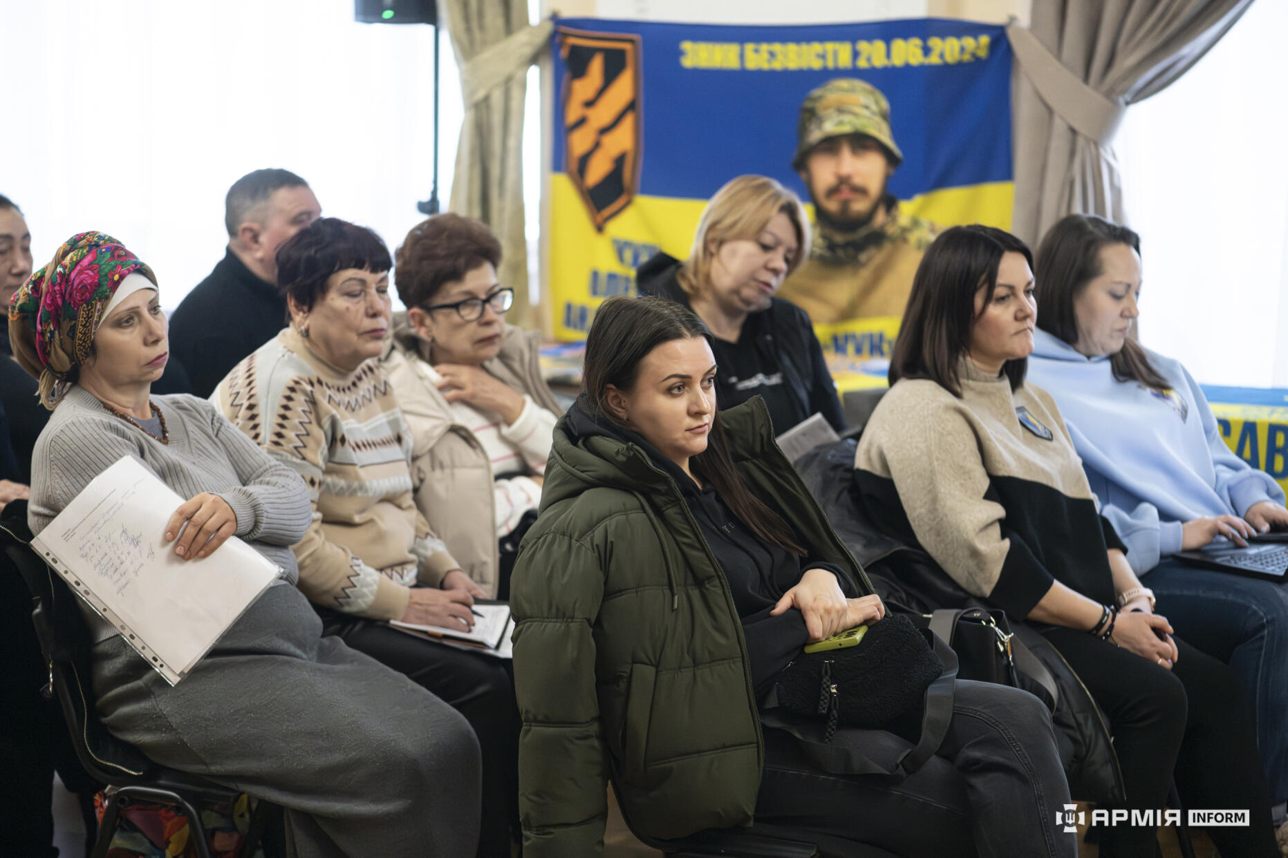 У першому ряду сидять жінки, які уважно слухають виступ під час зустрічі. Позаду них видно банер із датою та символікою військового підрозділу.