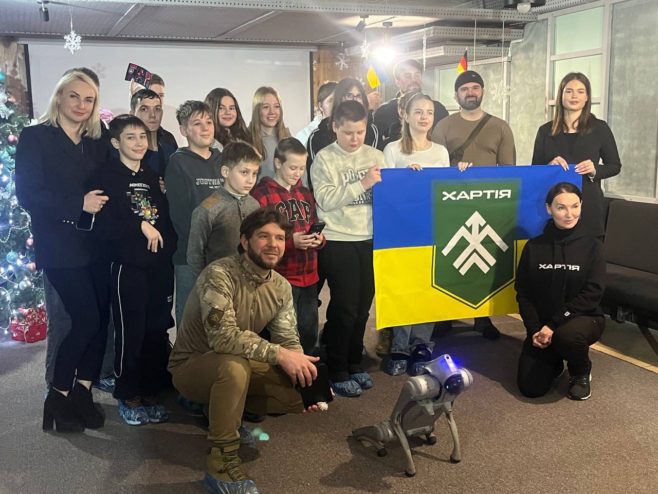 Велика група дітей та дорослих зібралася для спільного фото поруч із новорічною ялинкою. У центрі вони тримають прапор із символікою «ХАРТІЯ», а на підлозі перед ними стоїть робот-собака.