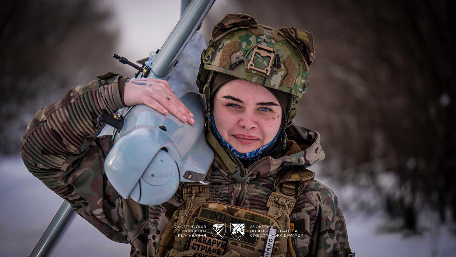 Жінка-військова у шоломі тримає великий безпілотник, дивлячись прямо в камеру. Її спокійний вираз обличчя поєднується з сучасним технічним оснащенням.