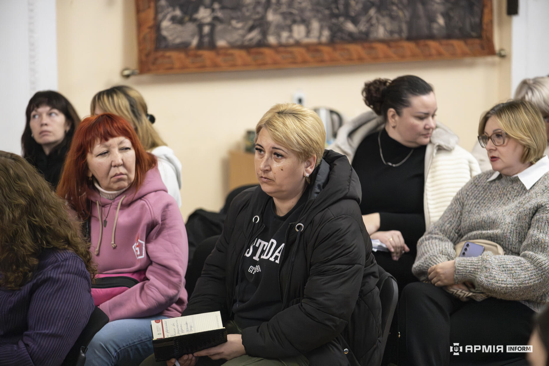 Група жінок під час обговорення сидить у залі, уважно слухаючи виступи та роблячи нотатки. Деякі з них тримають у руках блокноти або телефони, фіксуючи важливу інформацію.