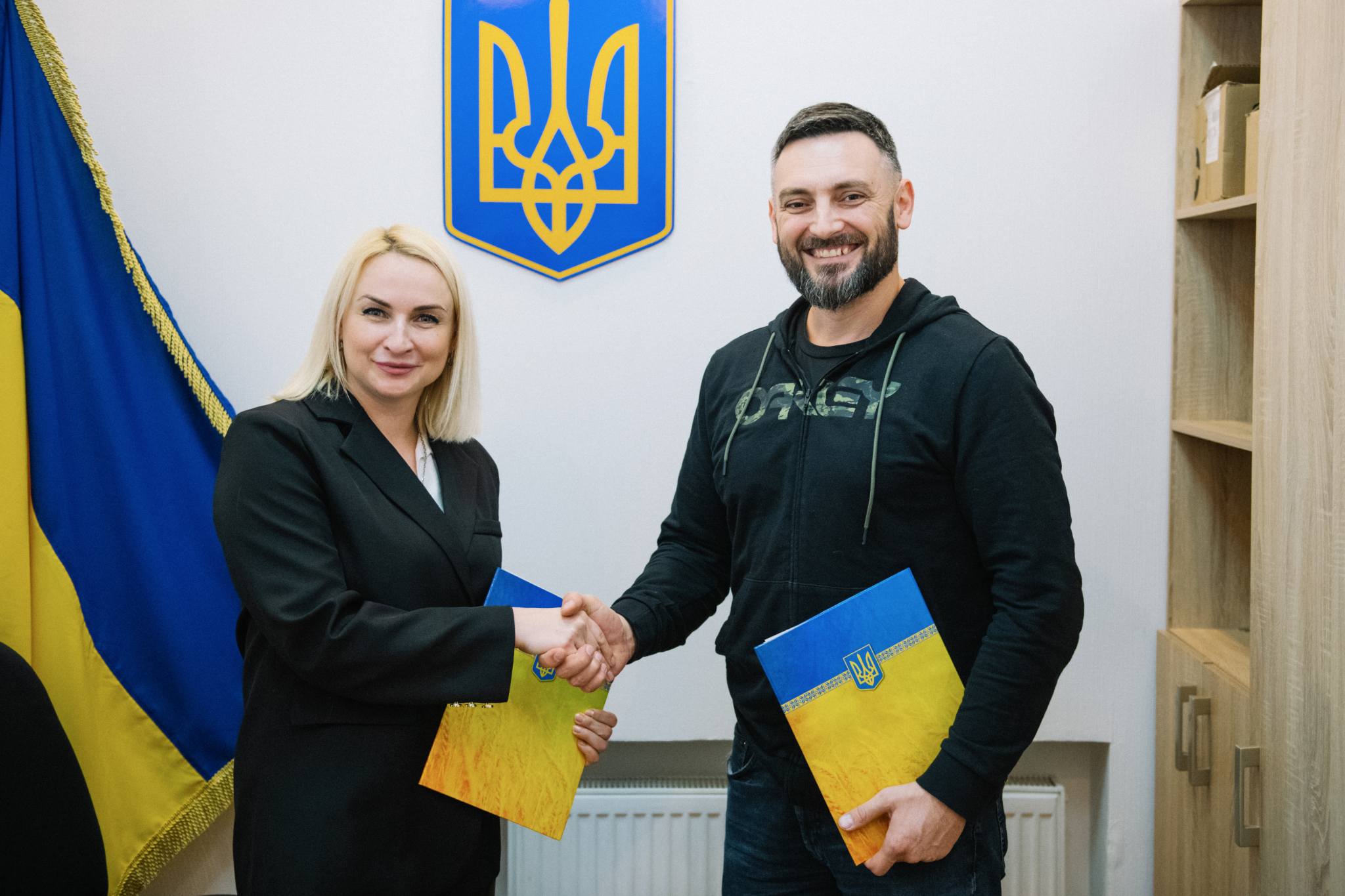 Жінка та чоловік тиснуть одне одному руки після підписання документів. У руках вони тримають папки з українською символікою, стоячи на тлі державного герба та прапора України.