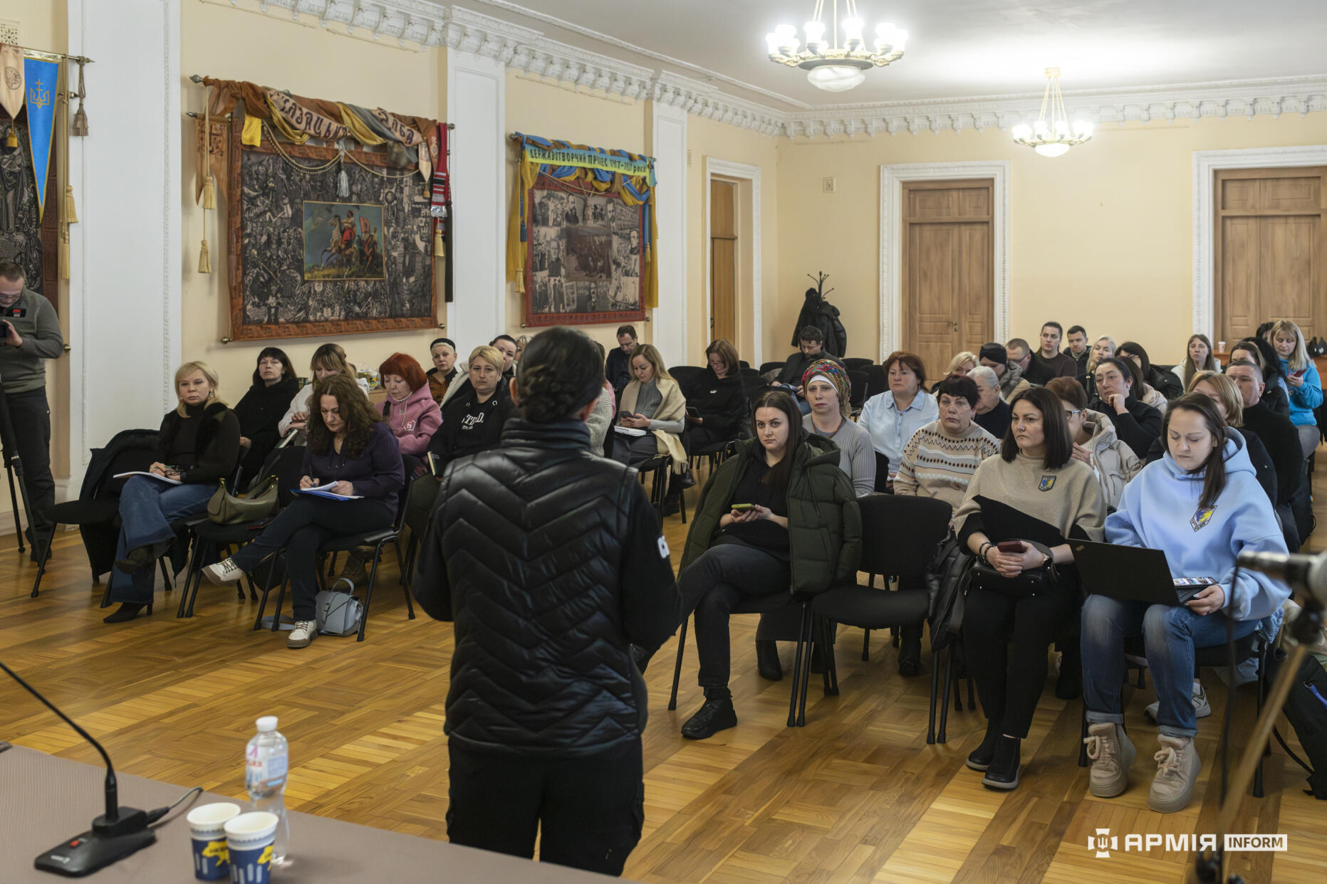 У просторій залі проходить зустріч, під час якої перед аудиторією виступає спікерка. Учасники сидять рядами, уважно слухаючи та роблячи нотатки.