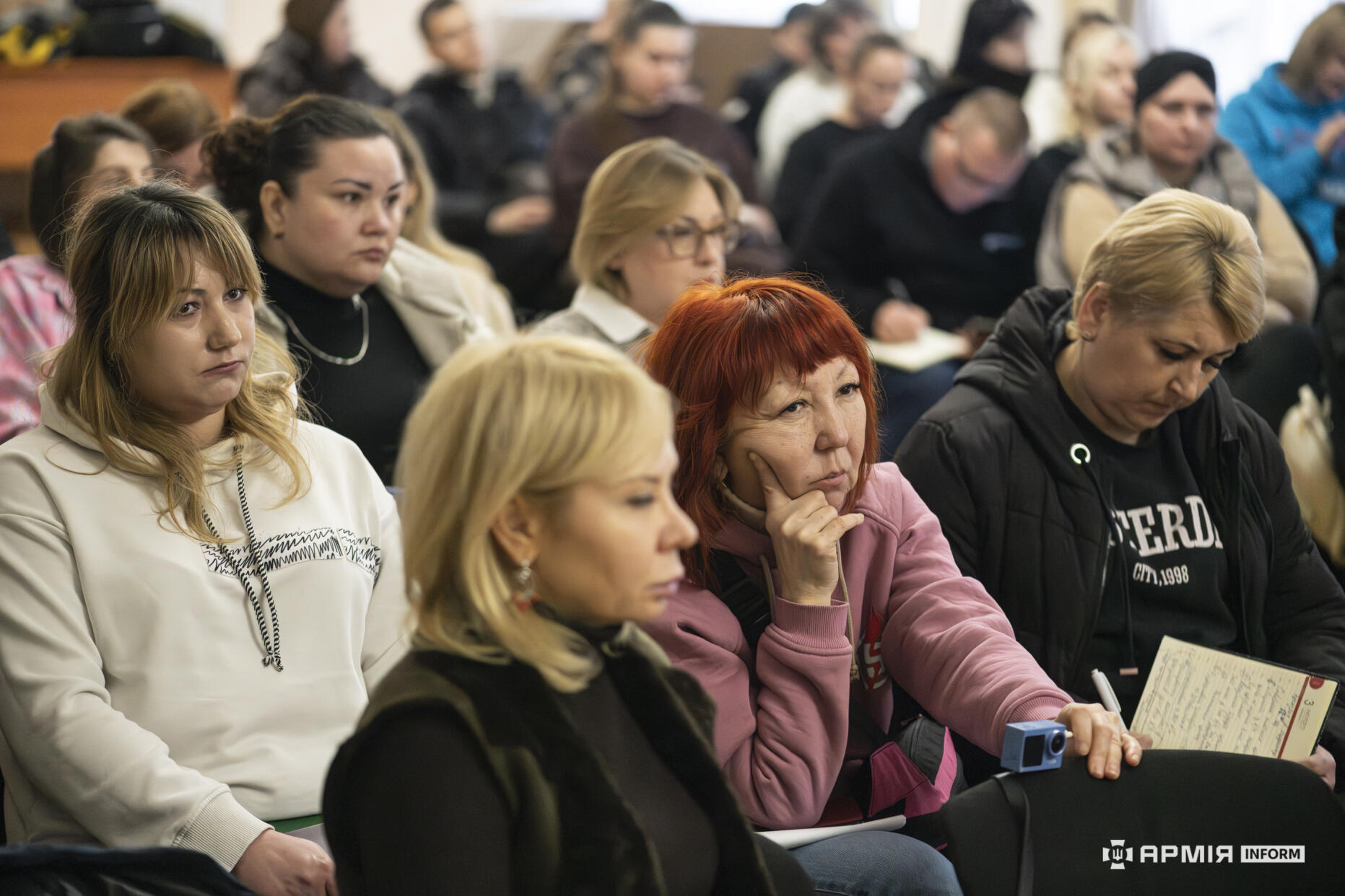 Кілька учасниць заходу зосереджено слухають виступ, одна з них робить записи у блокноті. Їхні погляди спрямовані вперед, де триває обговорення.