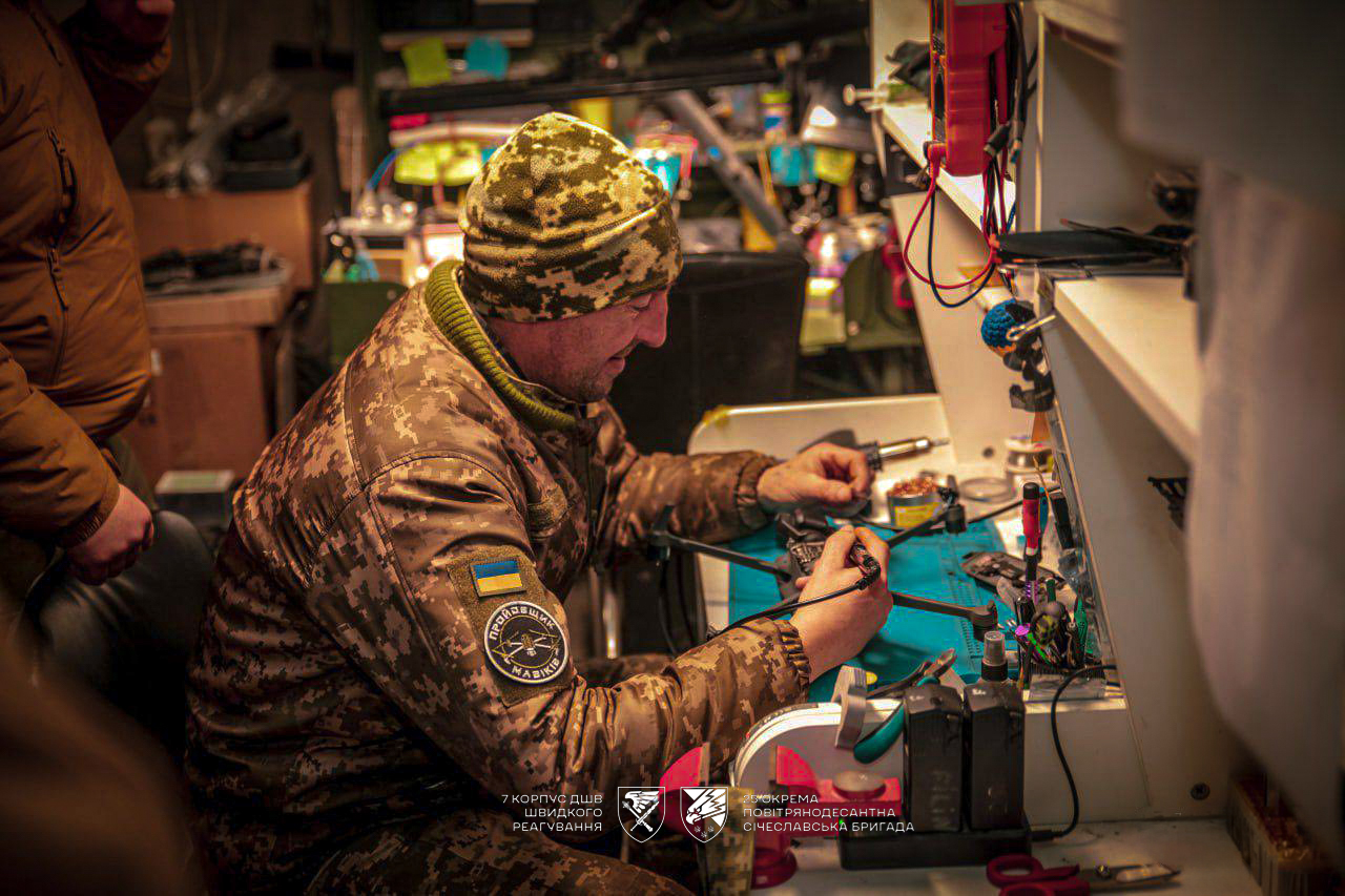 Військовий у майстерні зосереджено працює з електронікою, тримаючи паяльник біля деталей. Навколо нього — інструменти та компоненти, необхідні для ремонту техніки.
