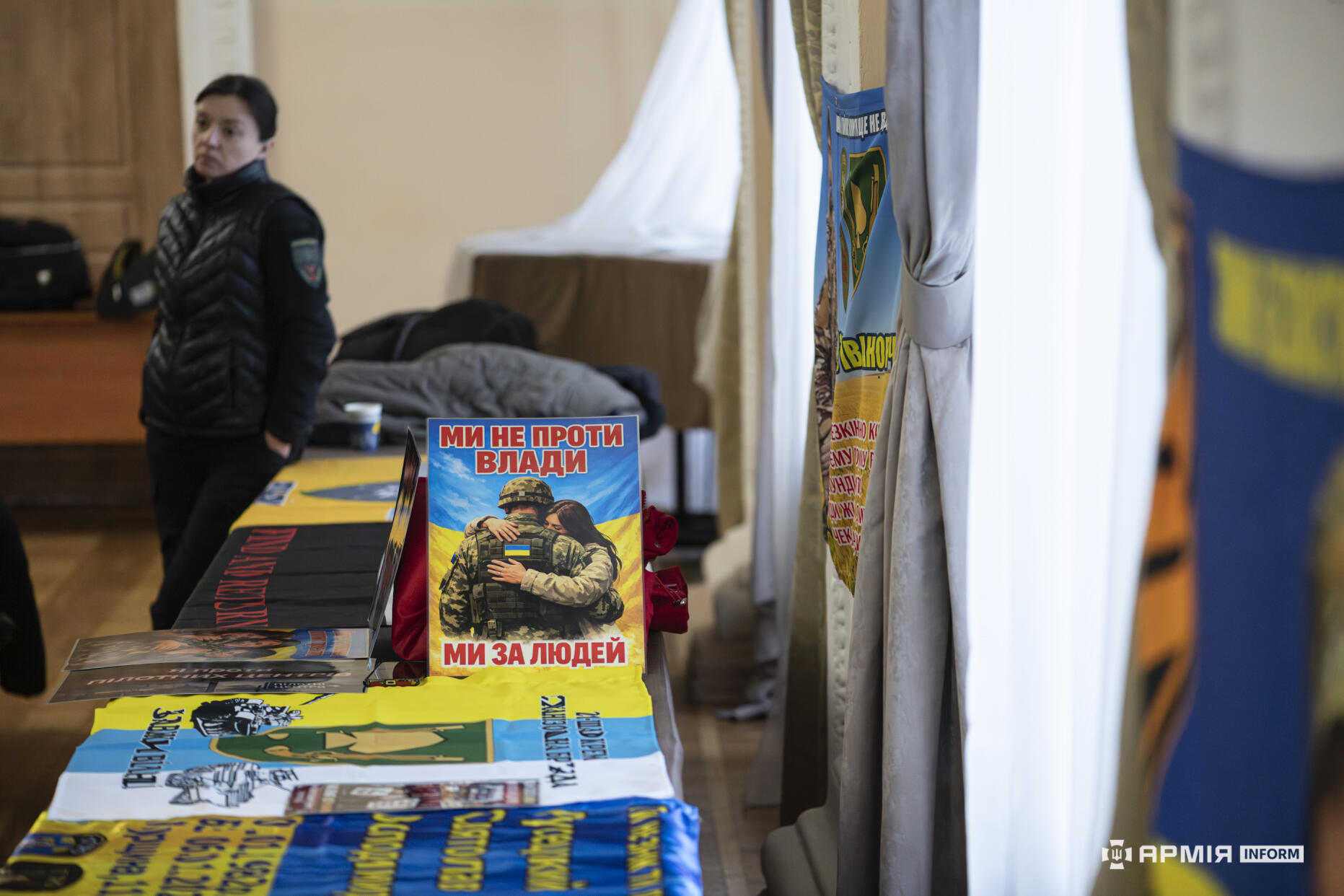На столі розкладені прапори та інформаційні матеріали, пов’язані з військовим підрозділом. У центрі стоїть плакат із написом «Ми не проти влади — ми за людей» і зображенням військового та цивільної.