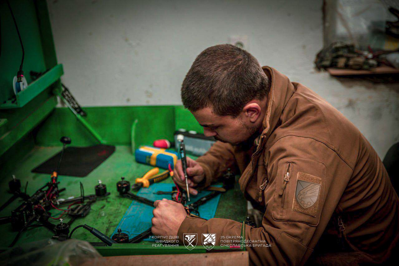 Інший боєць у робочому приміщенні займається складанням або ремонтом дрона, уважно обробляючи деталі. Робочий стіл заповнений інструментами та комплектуючими.