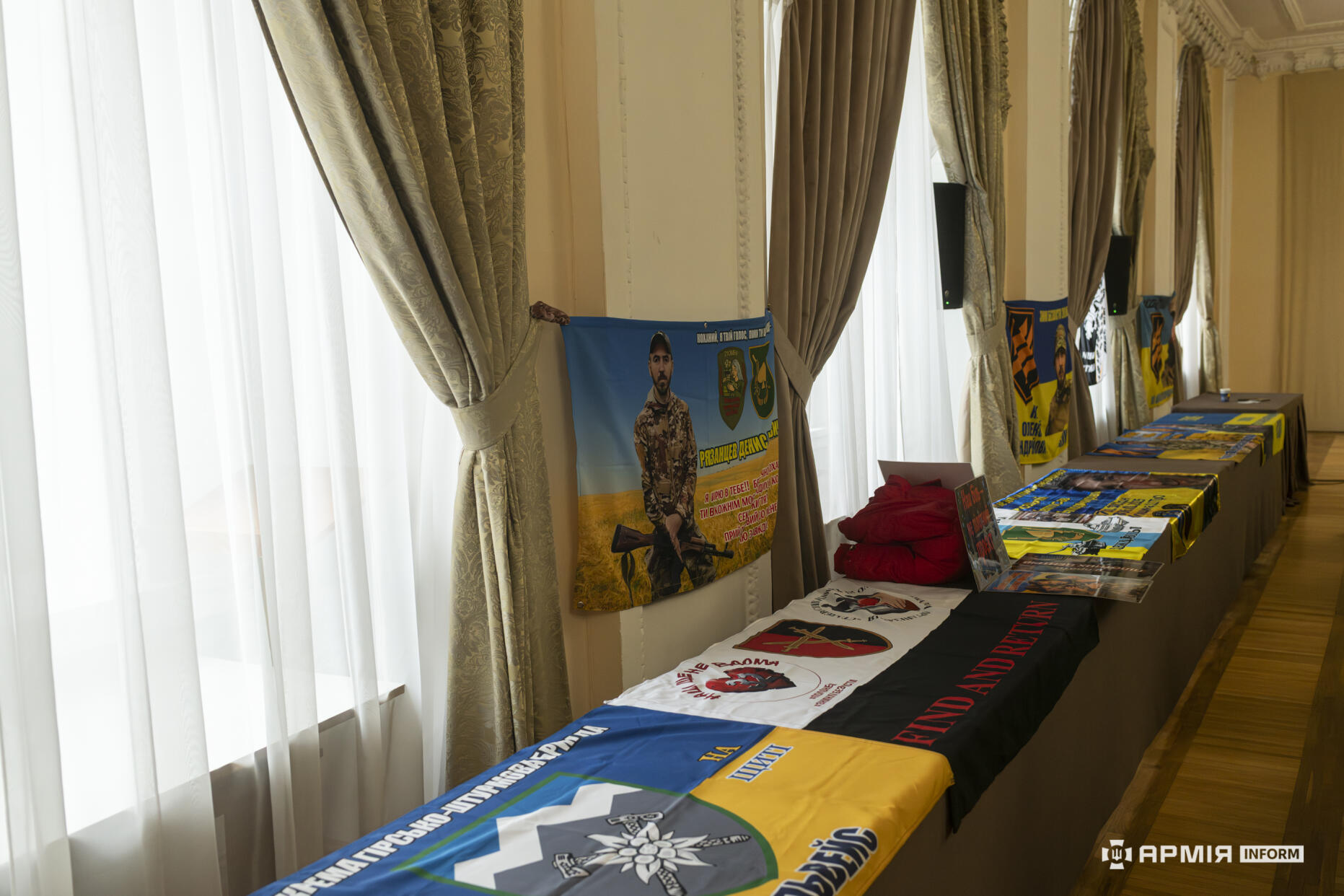 Уздовж стіни в залі розкладені прапори підрозділів і тематичні матеріали, присвячені українським військовим. Поруч на стіні розміщено банер із зображенням бійця та символікою підрозділу.