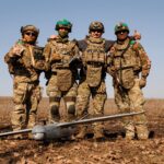 Четверо військових у повному спорядженні стоять разом у полі, обійнявшись за плечі та демонструючи позитивний настрій. Перед ними на землі лежить безпілотник літакового типу, що свідчить про їхню спільну роботу з дронами.