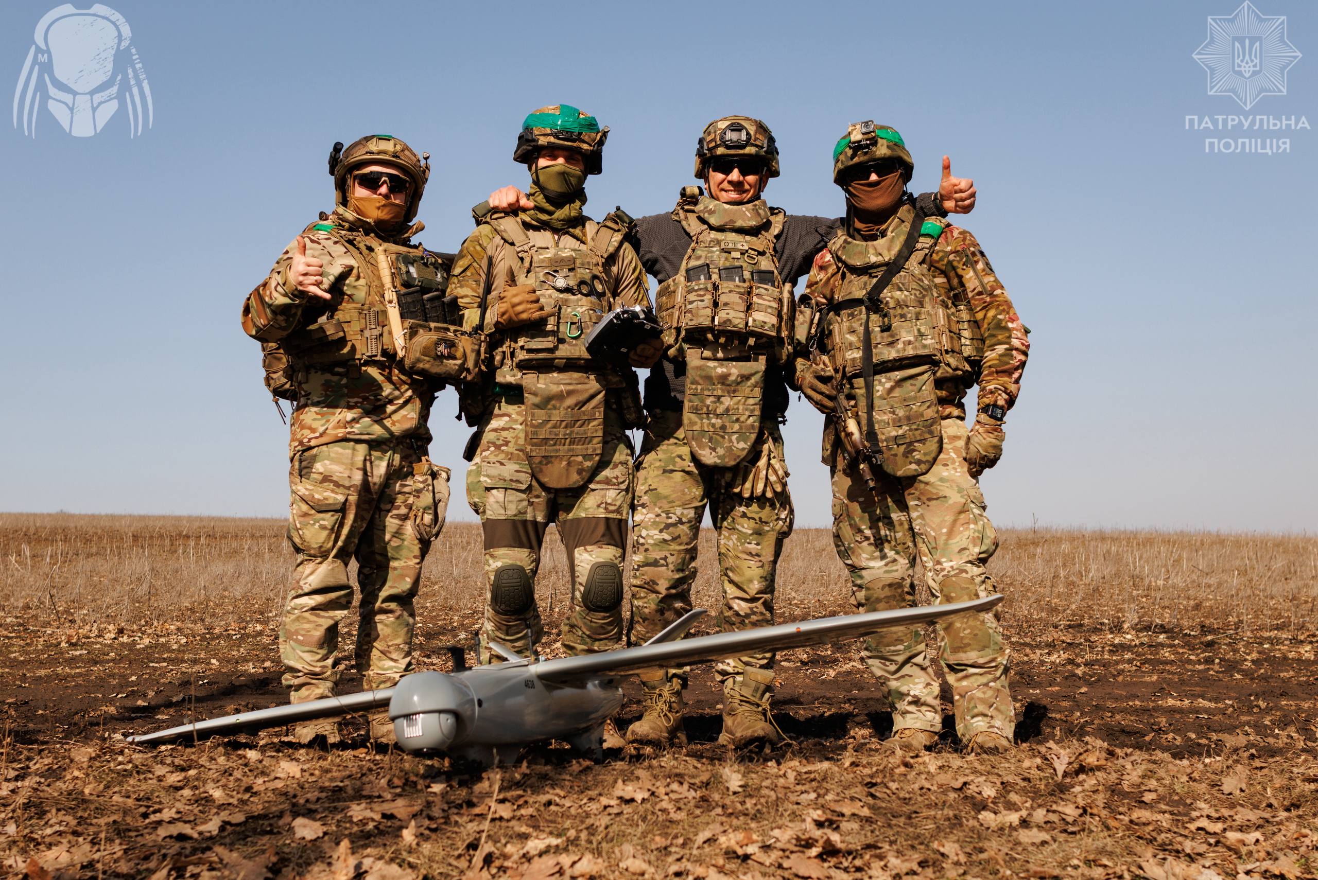 Четверо військових у повному спорядженні стоять разом у полі, обійнявшись за плечі та демонструючи позитивний настрій. Перед ними на землі лежить безпілотник літакового типу, що свідчить про їхню спільну роботу з дронами.
