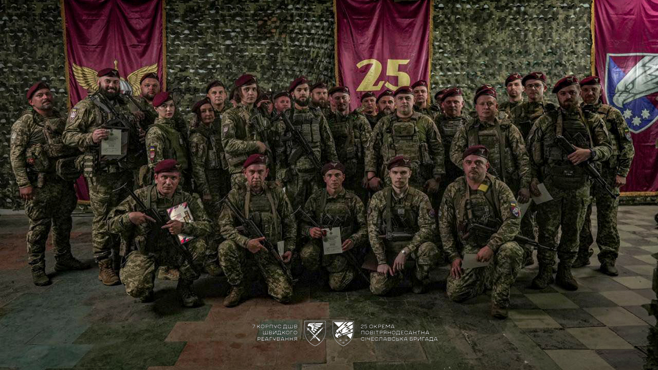 Велика група військових у беретах позує для спільного фото, тримаючи документи та зброю. На задньому плані видно прапори та символіку, що підкреслюють належність до бригади.