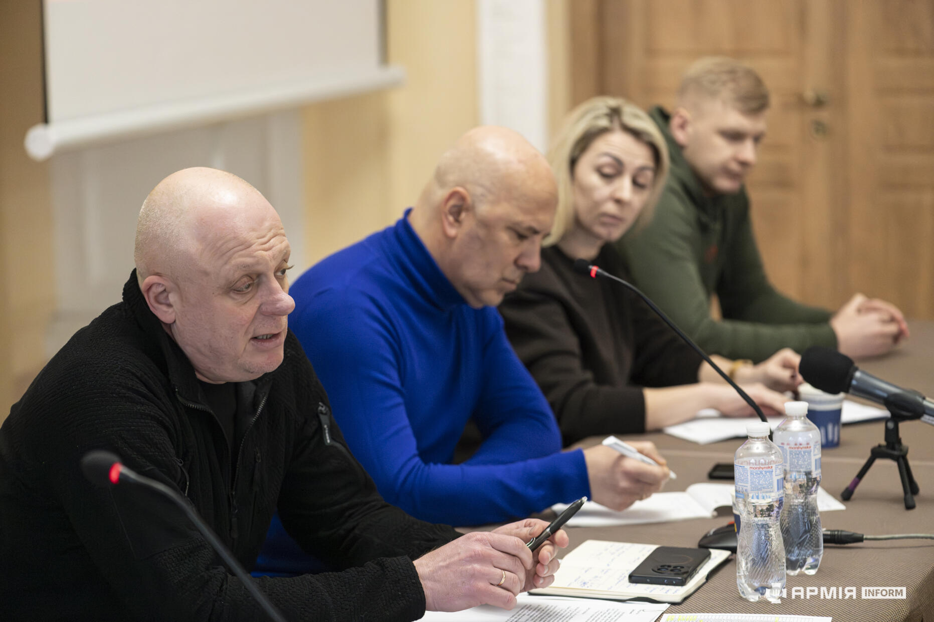 Учасники панелі сидять за столом під час зустрічі та виступають перед аудиторією. Один зі спікерів говорить у мікрофон, тоді як інші роблять нотатки або уважно слухають.