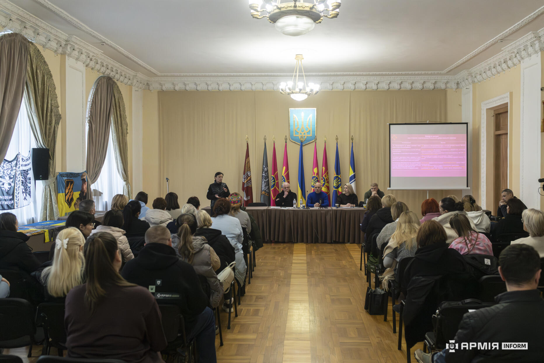 У просторій залі відбувається зустріч із представниками військових структур та родинами військовослужбовців. Перед аудиторією розташований стіл спікерів, позаду якого встановлені прапори та державний герб України.