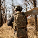 Боєць іде лісосмугою, несучи на плечі безпілотник, готовий до розгортання. На його шоломі закріплене обладнання, а на спині — рюкзак із спорядженням.