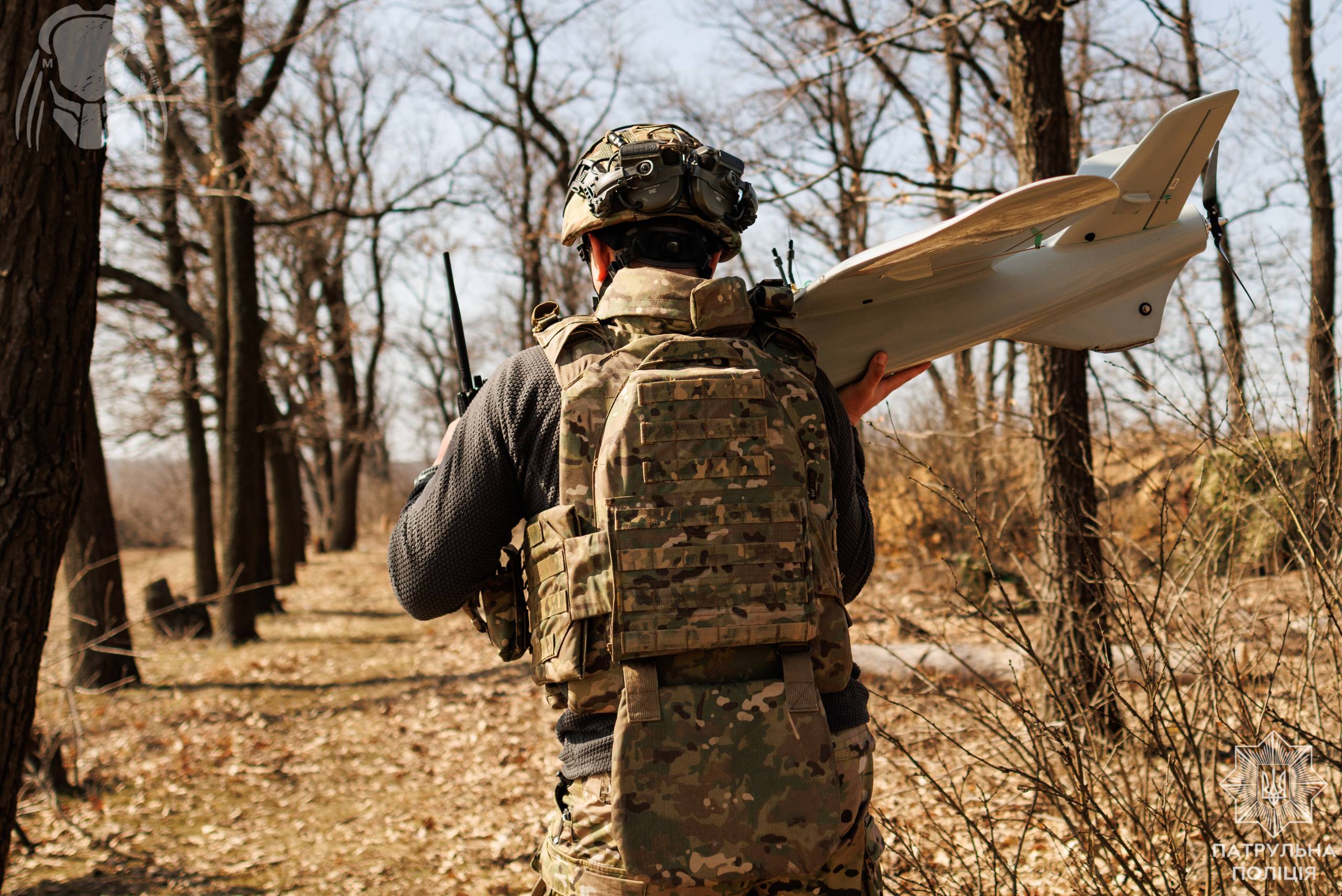 Боєць іде лісосмугою, несучи на плечі безпілотник, готовий до розгортання. На його шоломі закріплене обладнання, а на спині — рюкзак із спорядженням.