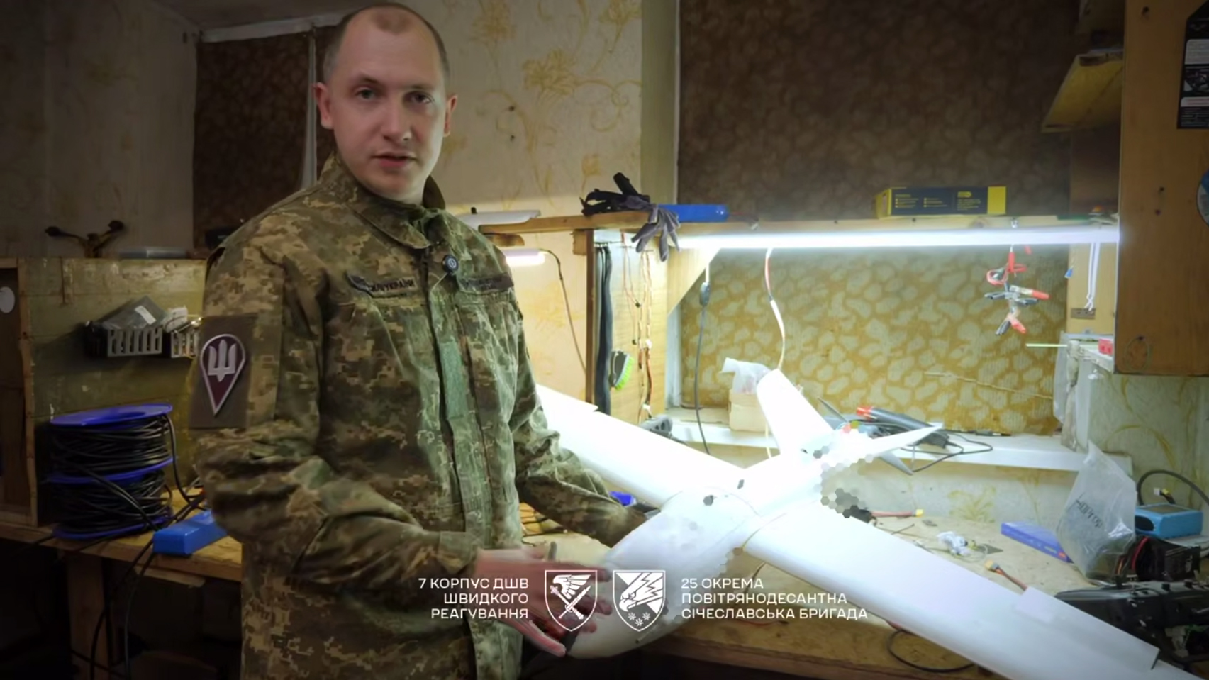 Військовослужбовець у формі стоїть у майстерні та тримає в руках безпілотник літакового типу, ймовірно під час налаштування або складання. На робочому столі позаду видно інструменти, освітлення та дроти, що свідчить про процес виготовлення або ремонту техніки.