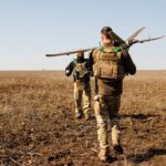 Двоє військових рухаються відкритим полем, один несе безпілотник, інший тримає апаратуру керування. Суха земля та відсутність укриття підкреслюють польові умови виконання завдання.