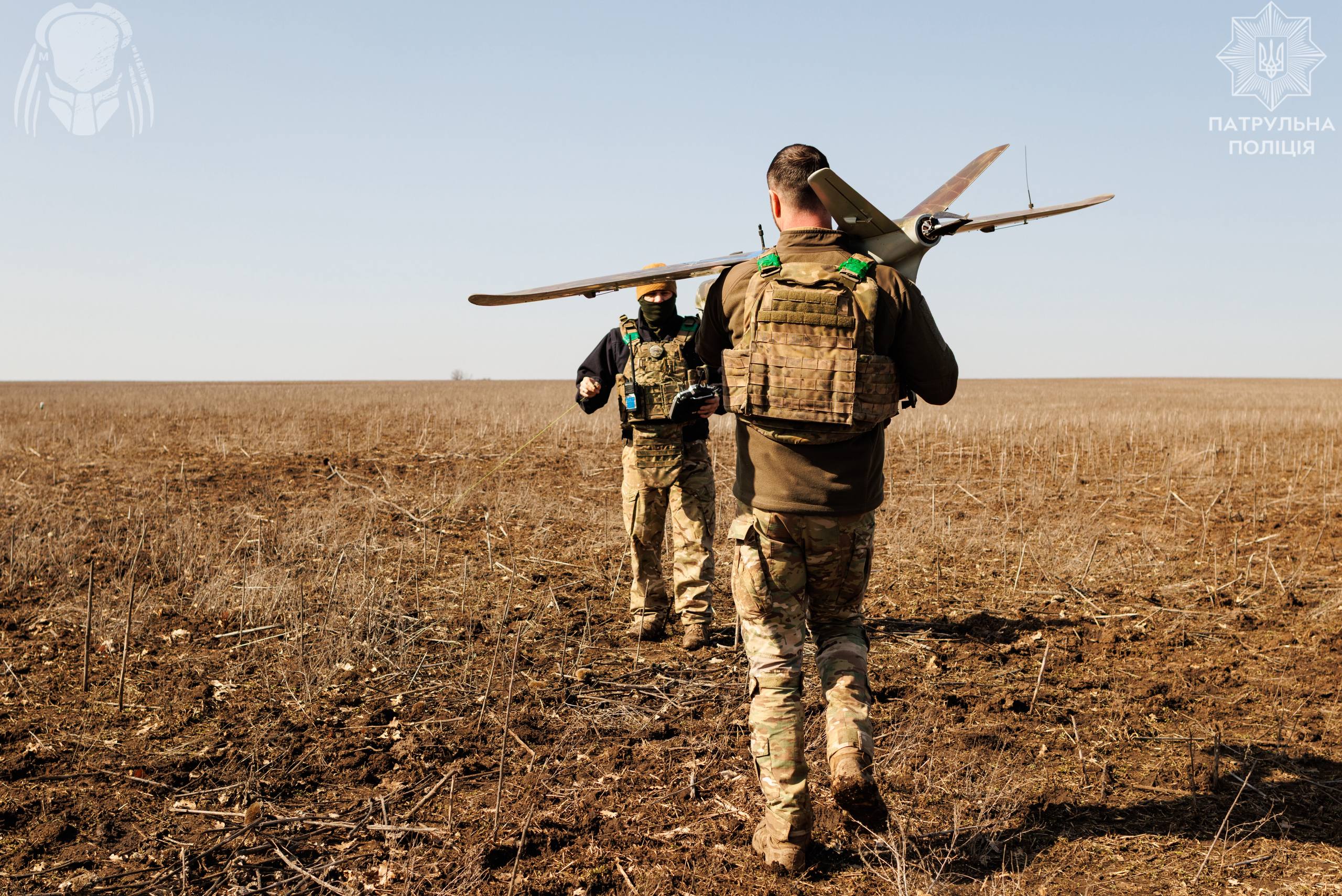 Двоє військових рухаються відкритим полем, один несе безпілотник, інший тримає апаратуру керування. Суха земля та відсутність укриття підкреслюють польові умови виконання завдання.