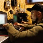 Оператор сидить у невеликому приміщенні та працює за ноутбуком, зосереджено дивлячись на екран. Навколо нього розміщене обладнання для керування або налаштування техніки.