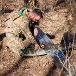 Військовий стоїть на колінах у лісі та налаштовує безпілотник, відкривши його корпус. Поруч видно замасковану позицію, що вказує на підготовку до запуску.