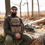 Військовий сидить на замаскованій позиції, поруч із ним лежить безпілотник. Його спокійна поза та впевнений вигляд контрастують із бойовими умовами навколо.