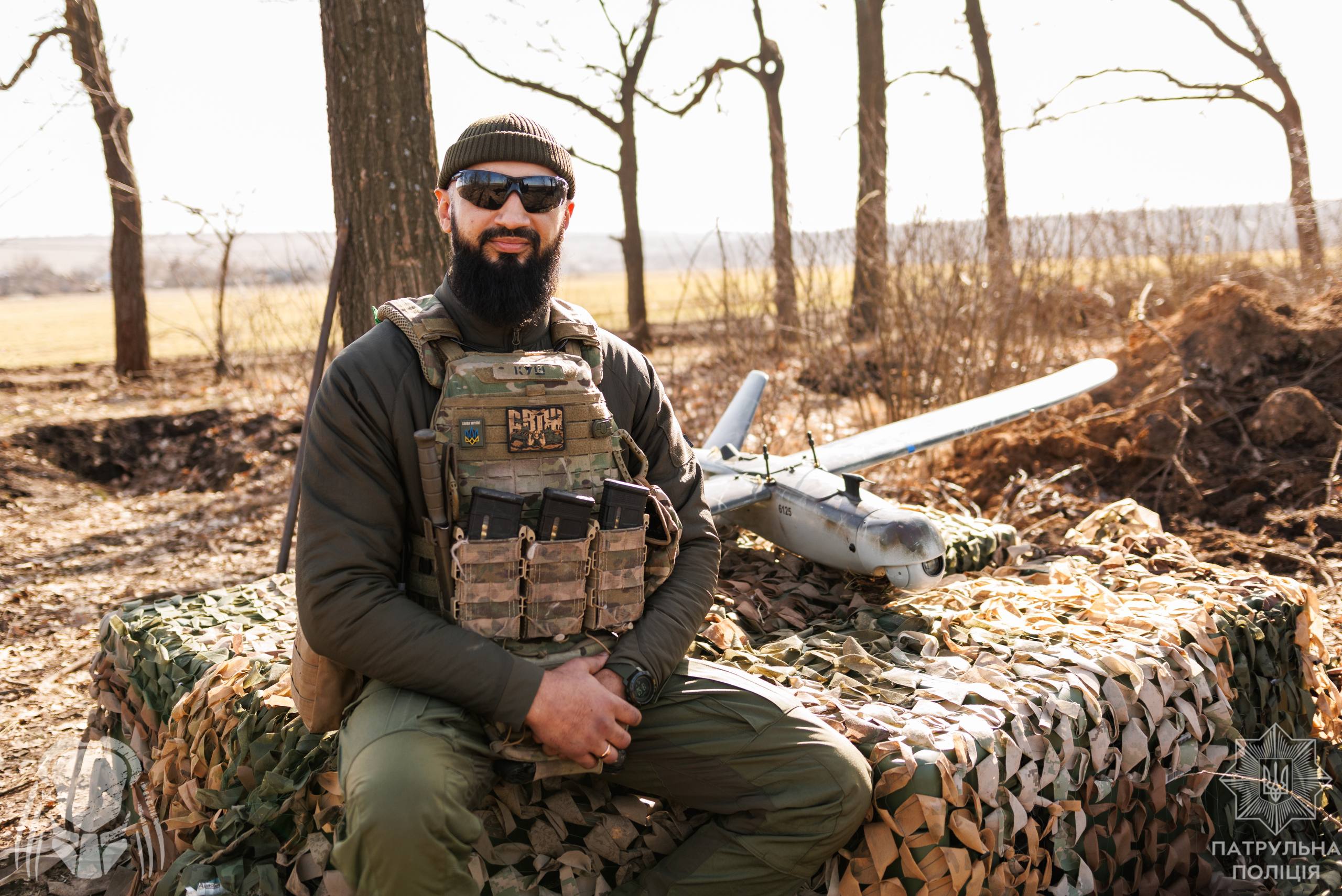 Військовий сидить на замаскованій позиції, поруч із ним лежить безпілотник. Його спокійна поза та впевнений вигляд контрастують із бойовими умовами навколо.