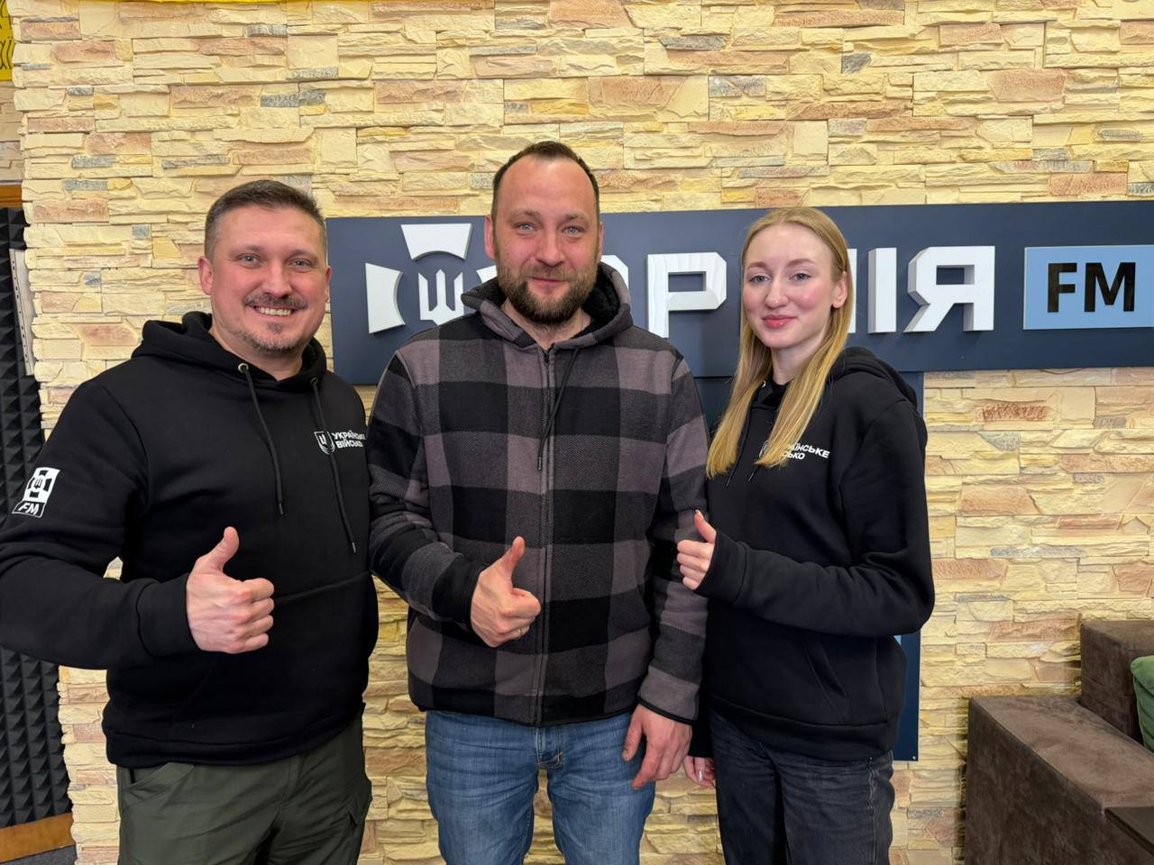 Двоє чоловіків та жінка стоять на тлі емблеми АрміяFM