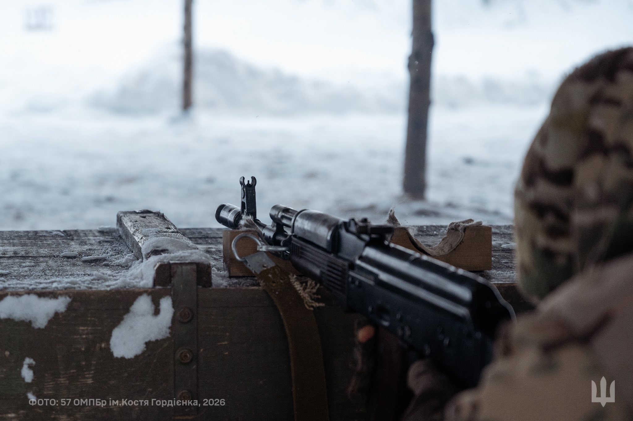 Боєць веде спостереження з укриття, тримаючи автомат, встановлений на дерев’яному упорі позиції. Перед ним розгортається засніжене поле з поодинокими деревами, що підкреслює холодні зимові умови на фронті.