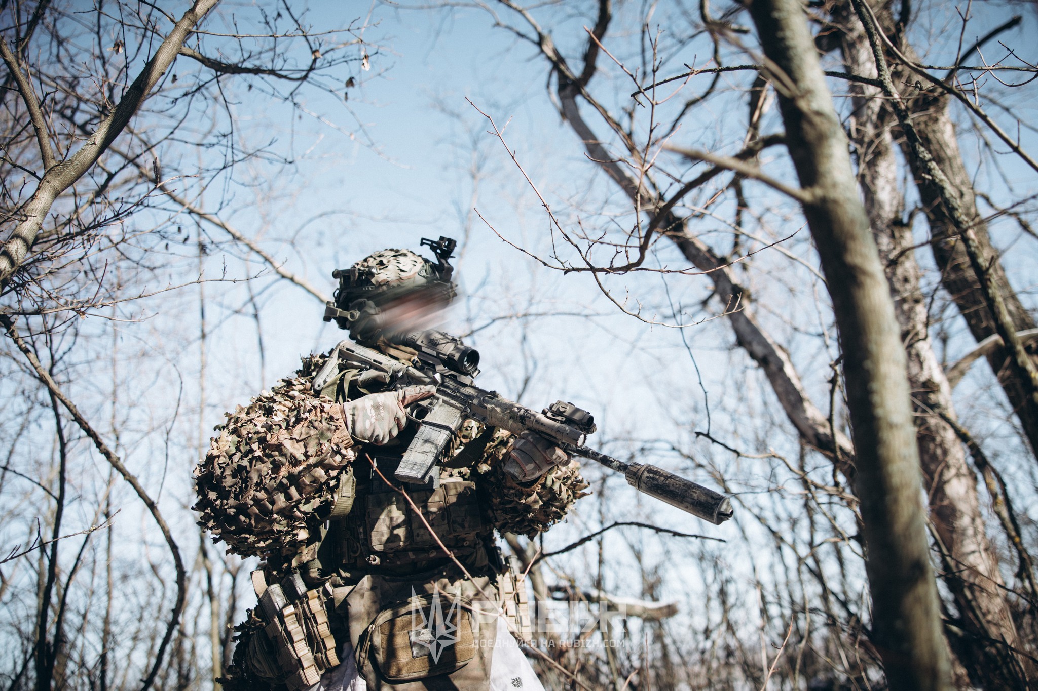 Військовий у маскувальному спорядженні пересувається серед дерев, тримаючи автомат із глушником напоготові. Його екіпірування та уважна постава свідчать про виконання завдання в умовах лісистої місцевості.